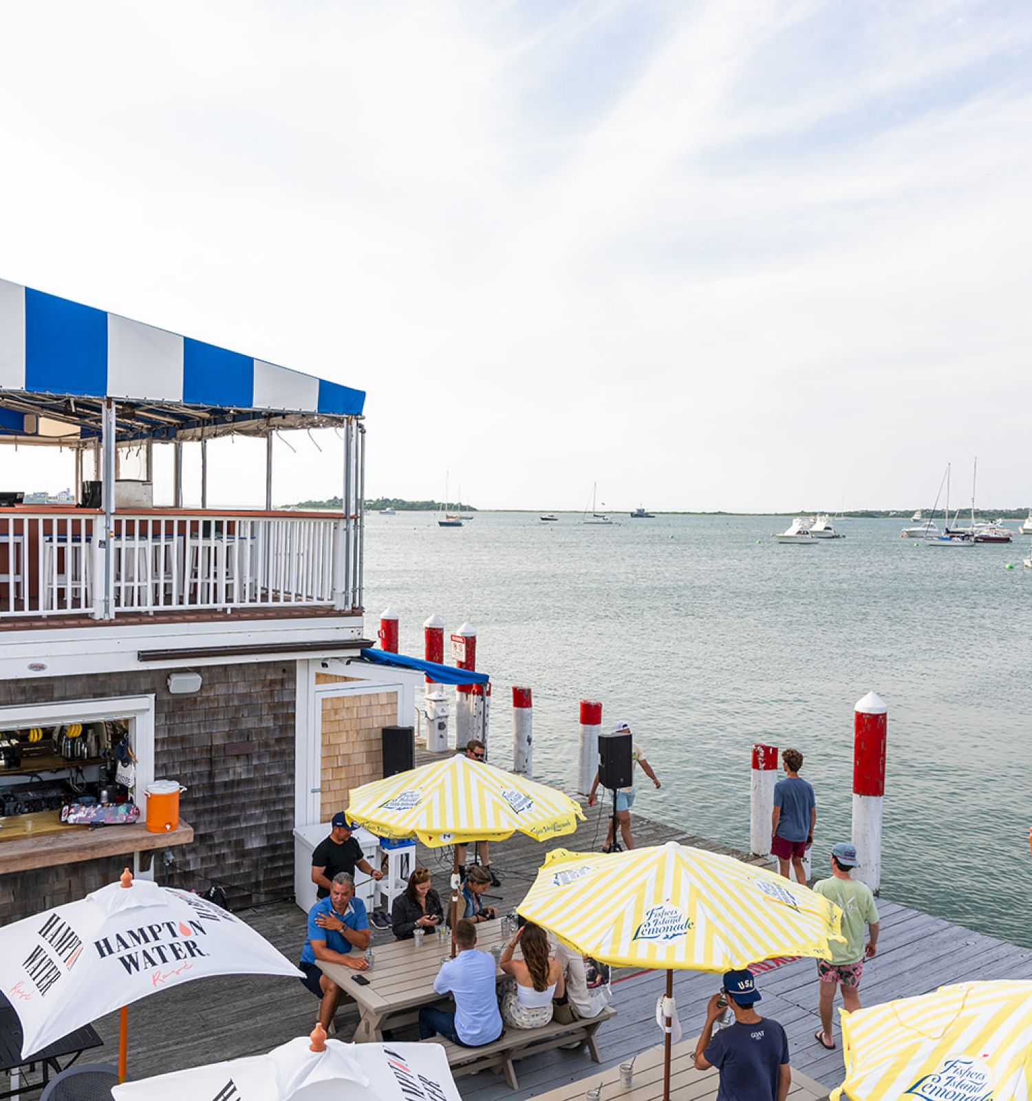 People are dining at an outdoor waterfront restaurant with yellow and white umbrellas, overlooking a marina with boats in the background.