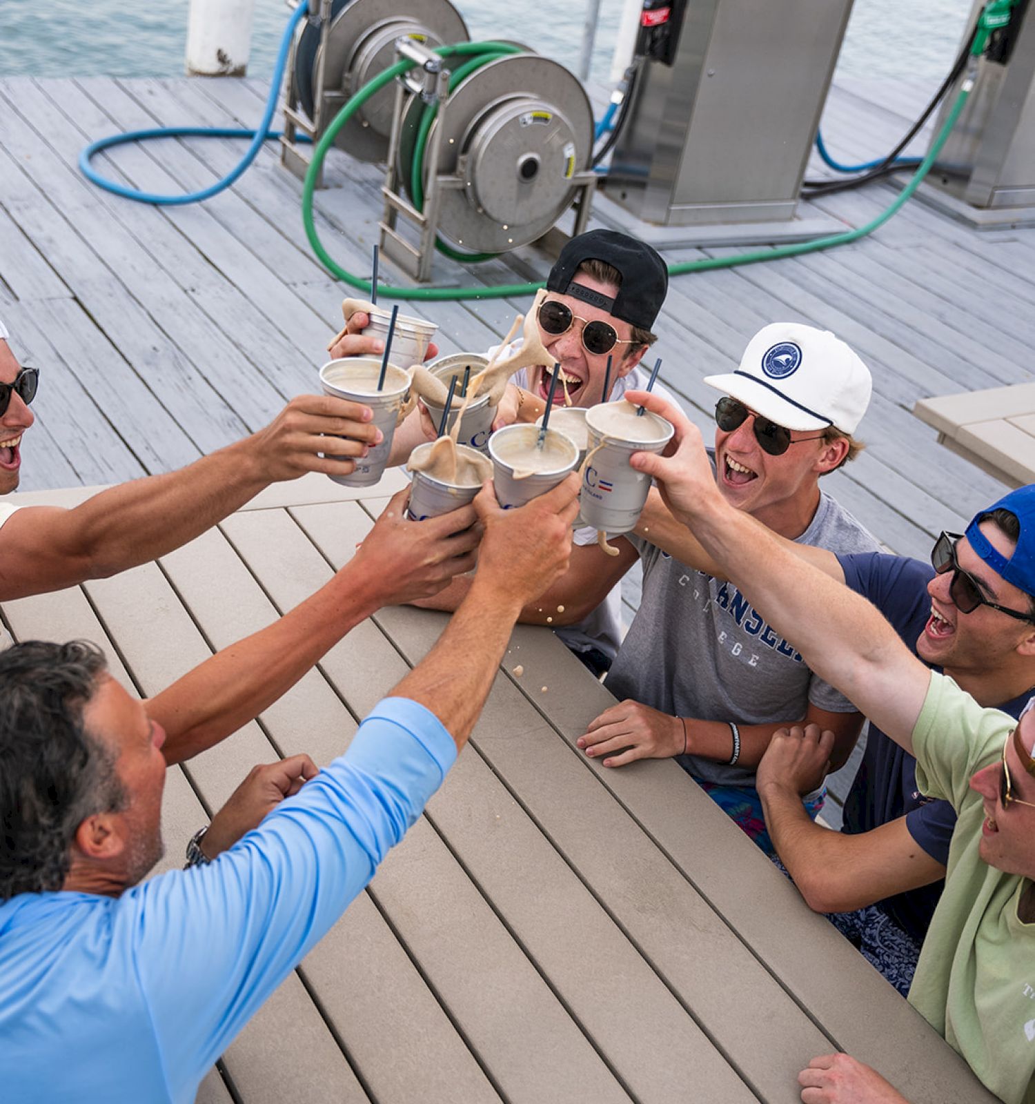 A group of people raising their drinks in a toast on a dock with fuel pumps and hoses in the background.