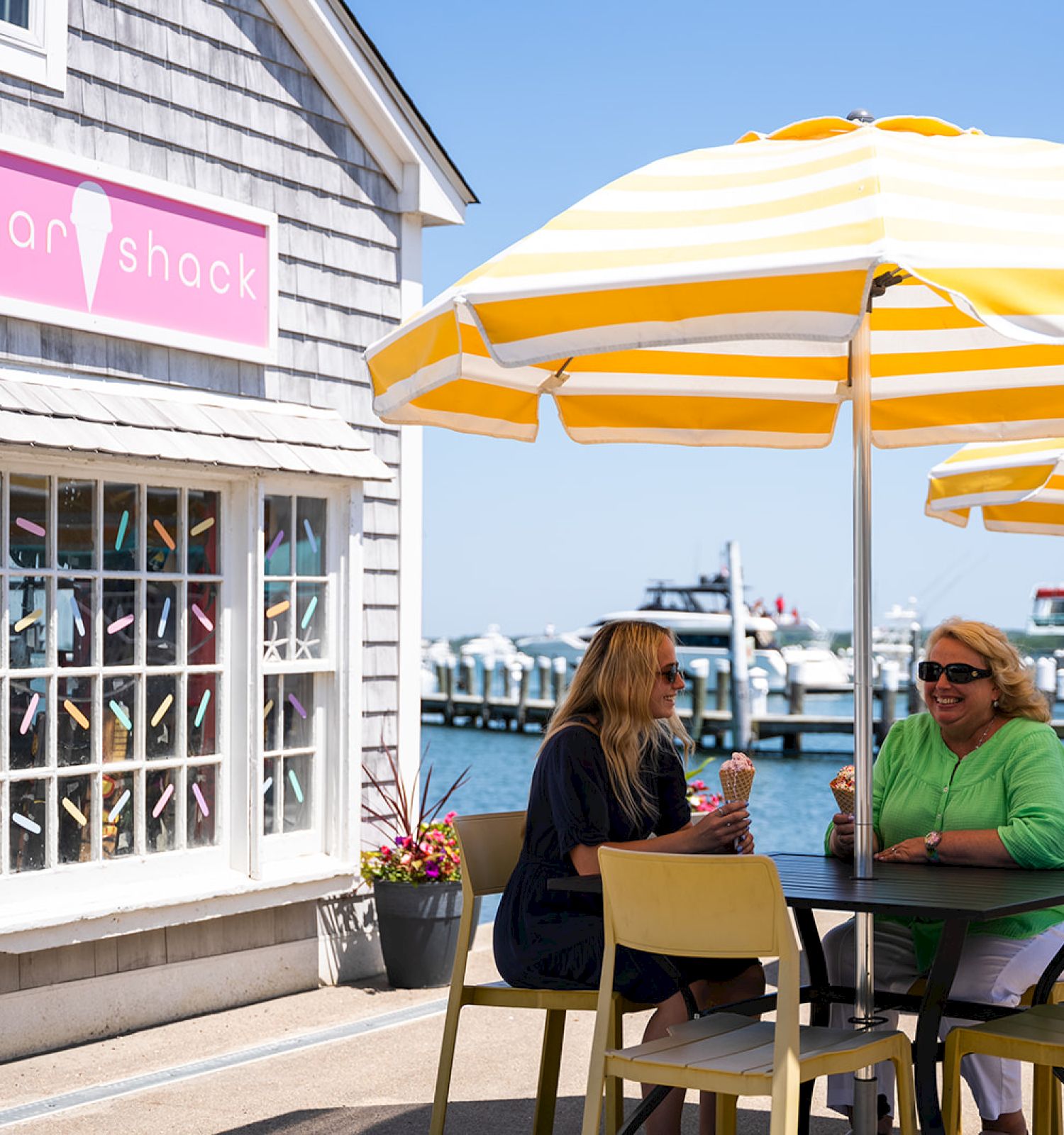 Two women are talking at an outdoor table under yellow and white umbrellas by a harbor, near a building that has a sign reading 