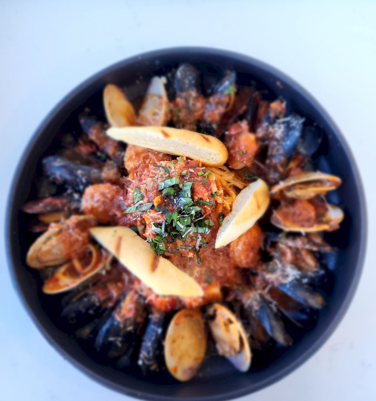 A black bowl filled with mussels, clams, and other seafood, garnished with herbs and sliced bread on top, all served in a savory sauce.