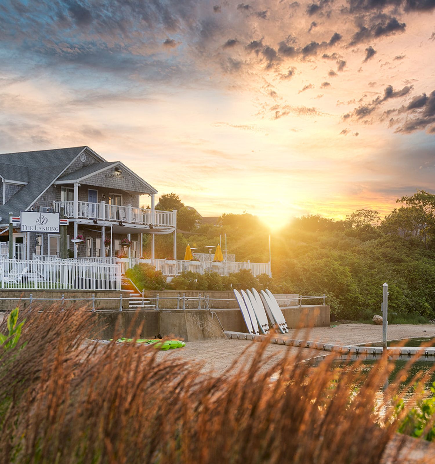 A house by the water with a deck, surrounded by greenery and flowers, with a beautiful sunset in the background ending the sentence.