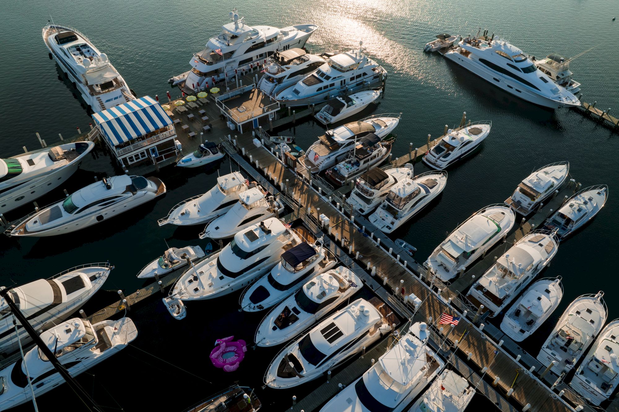 The image shows a marina with numerous yachts and boats moored at the docks, with sunlight reflecting off the water and vessels arranged closely.