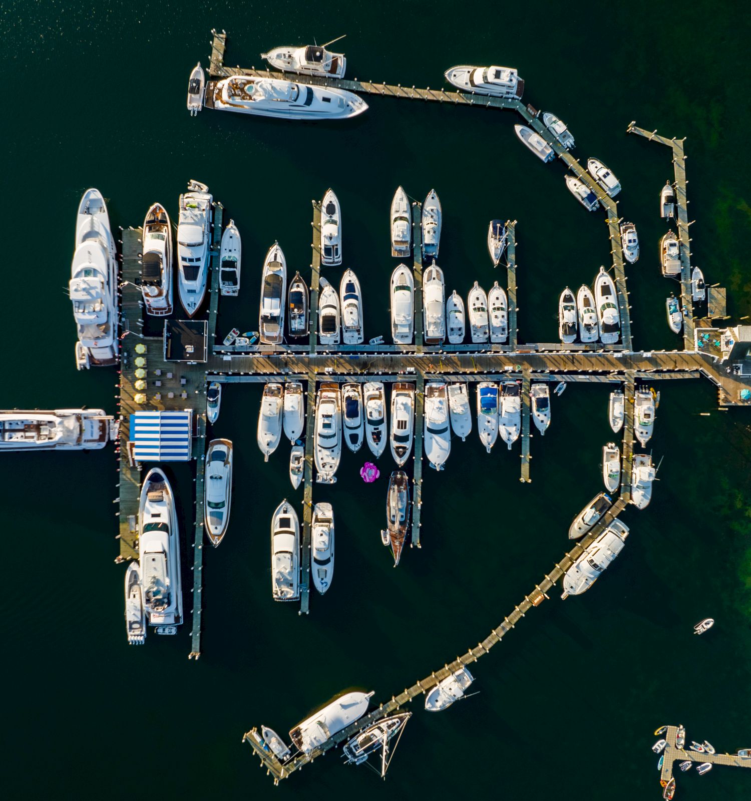 An aerial view of a marina filled with numerous boats and yachts docked along various piers, with a small building and vegetation visible on the right.