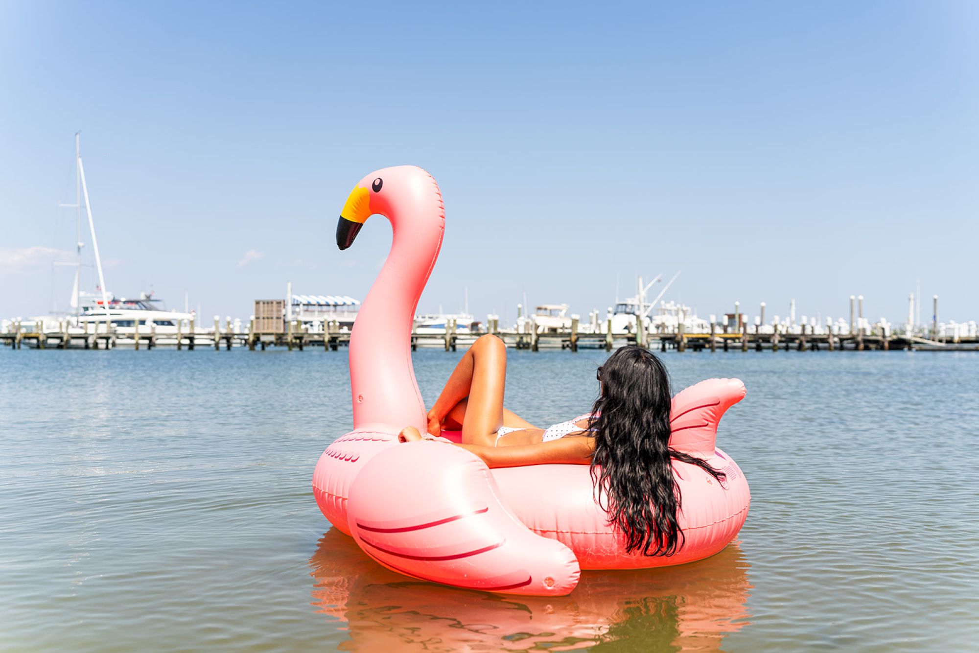 A person lounges on a large pink flamingo float in a calm body of water with boats docked in the background under a clear blue sky.
