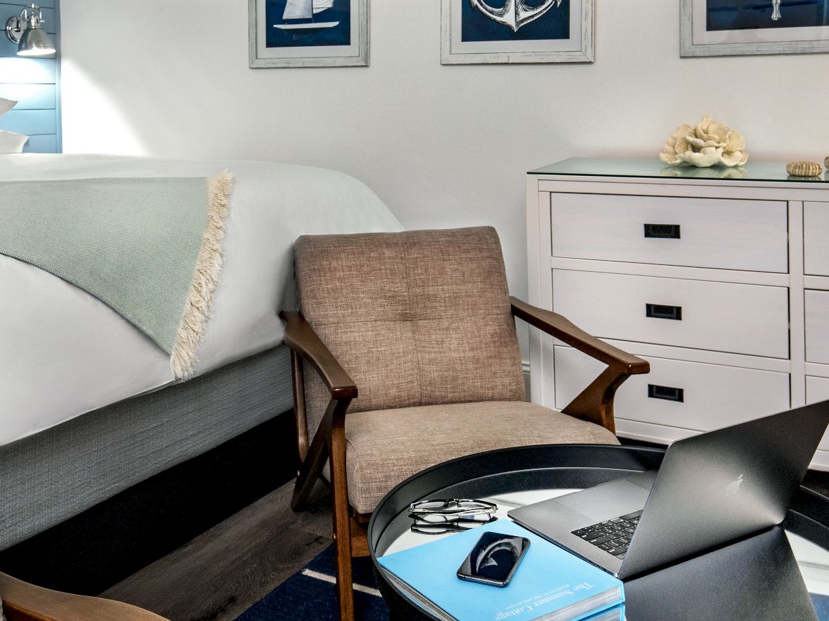 The image shows a cozy room with a bed, a chair, a dresser, a table with a laptop, a notebook, glasses, and a phone.