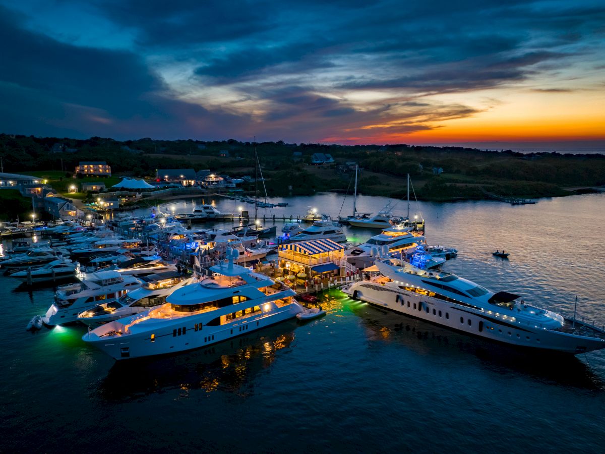An illuminated marina filled with yachts and boats, set against a vibrant sunset sky and coastal landscape.