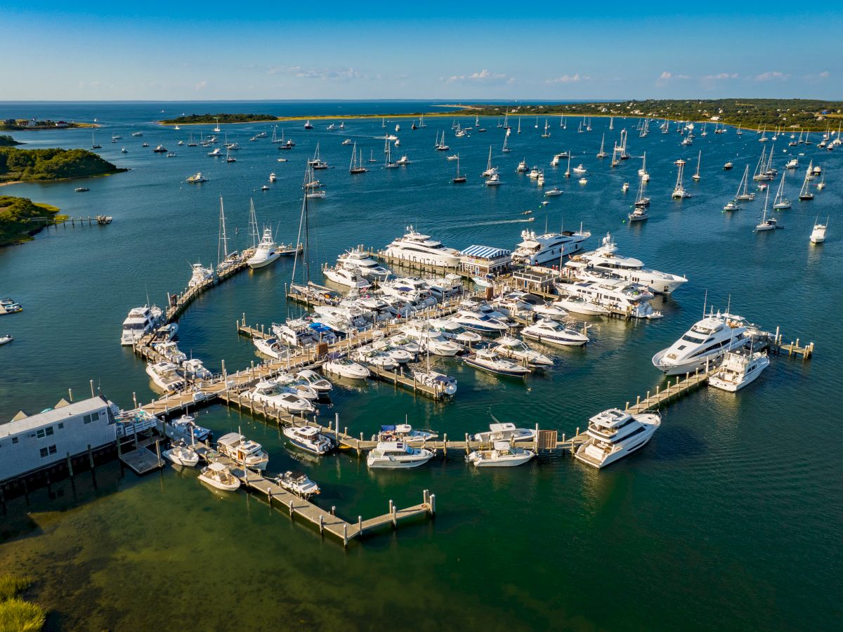 An aerial view of a marina filled with numerous boats and yachts, surrounded by calm waters and a scenic coastline extending into the distance.