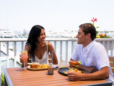Two people are dining outside, enjoying food and drinks on a sunny day by the waterfront, with boats visible in the background.