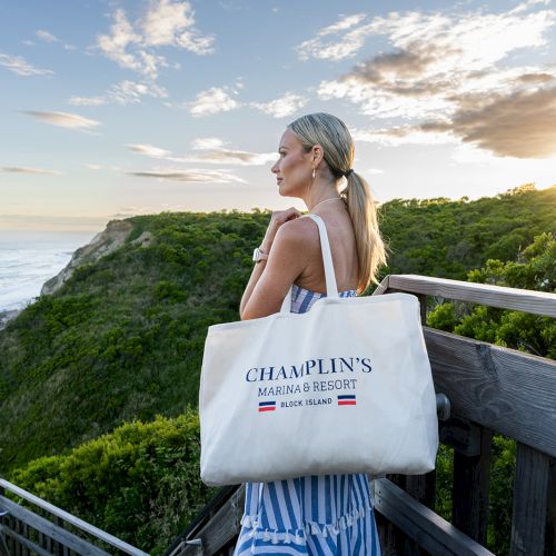 A woman stands on a boardwalk overlooking a scenic coastal view, holding a Champlin's Marina & Resort bag, with the sun setting in the background.