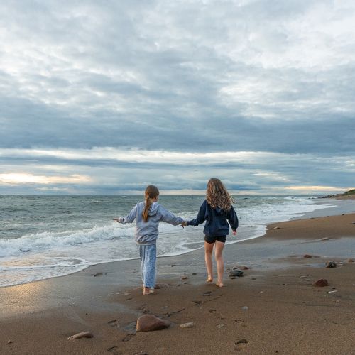 Two people walk hand in hand along a sandy beach with scattered rocks, near the ocean under a cloudy sky, creating a serene scene.