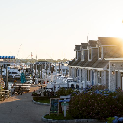 A quaint harbor scene with boats docked, cozy houses, outdoor seating areas, and the sun shining between the rooftops.