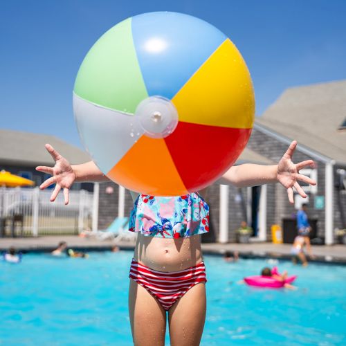 A child in a colorful swimsuit holds a beach ball over a swimming pool with people in the background, on a sunny day.