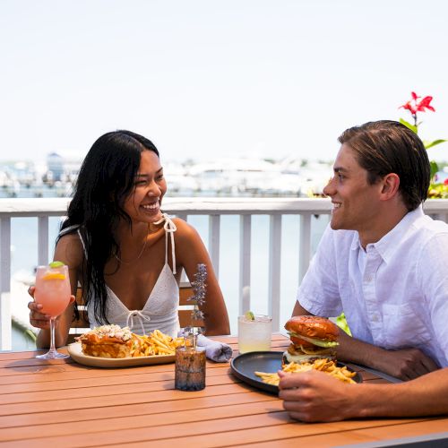 Two people are enjoying a meal outside on a sunny day, with a marina in the background. They are smiling and have food and drinks on the table.