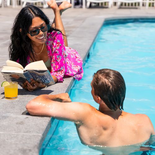 A woman in sunglasses reads a book while lounging by the poolside, and a man in the pool converses with her with a drink nearby.