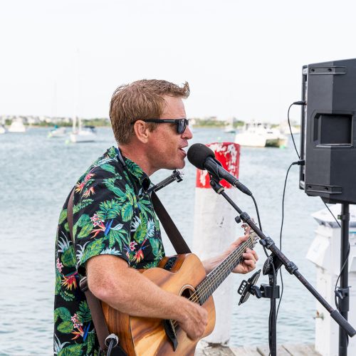 A man in a Hawaiian shirt plays a guitar and sings into a microphone on a dock, with water and boats visible in the background.