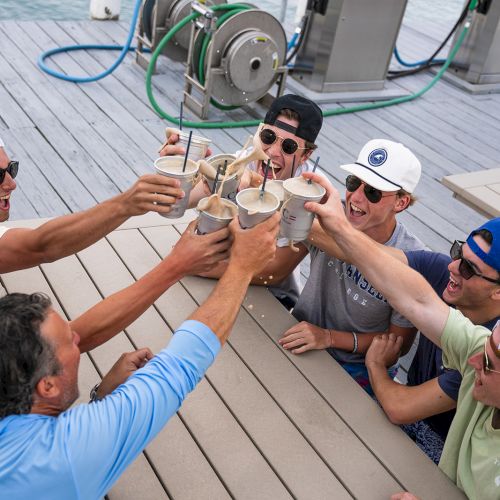 A group of people is toasting with drinks on a dock by the water, with equipment and hoses visible in the background.