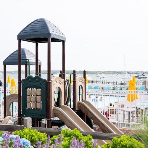 The image shows a playground with slides and other equipment near a pool, with yellow umbrellas and a body of water in the background.