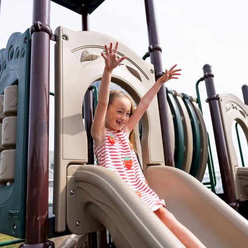 A child in a striped outfit joyfully slides down a playground slide with arms raised, enjoying a sunny day outdoor.