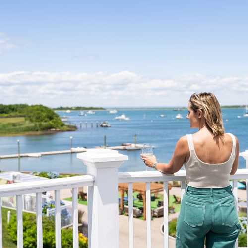 A woman stands on a balcony overlooking a scenic waterfront, holding a wine glass, enjoying a sunny day with boats and greenery in the distance.