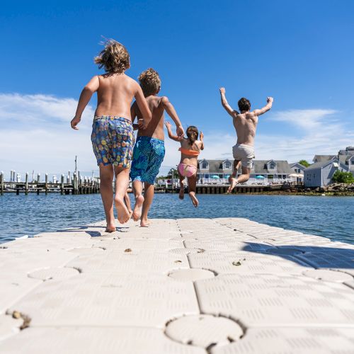Four children in swimsuits run and jump off a floating dock into a calm body of water, with buildings and a boat in the background.