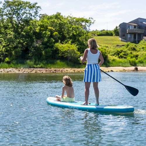 A person paddleboarding with a child on a calm body of water, surrounded by greenery and houses in the background, on a sunny day.