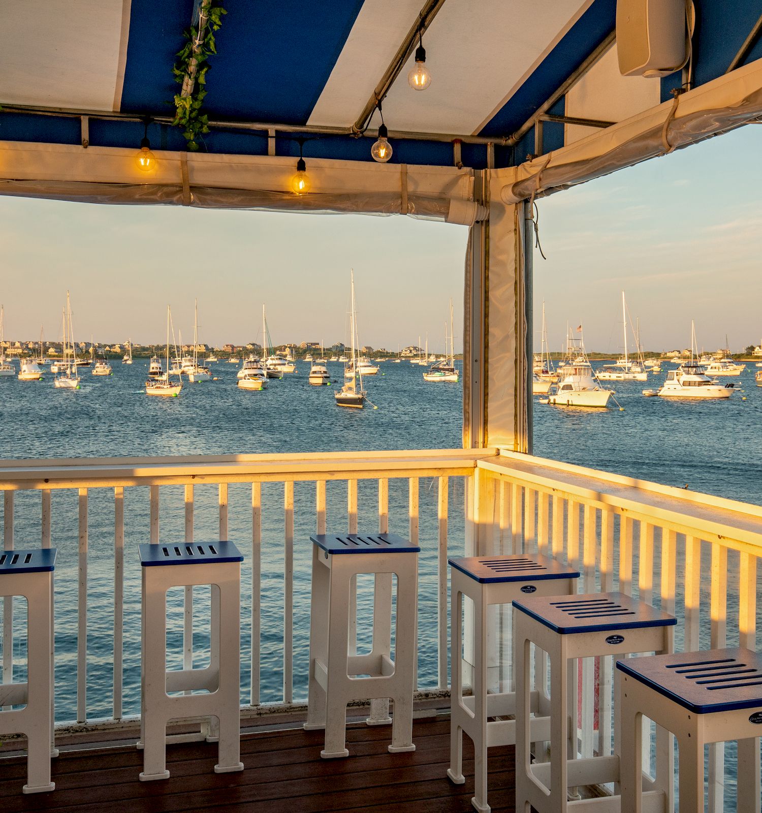 An outdoor seating area with white stools and a striped canopy overlooks a bay filled with sailboats on a sunny day, creating a serene atmosphere.