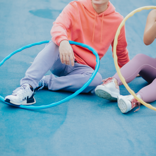 Two people sitting on a blue sports court, each holding a hula hoop, with one person smiling and the other mostly out of the frame.