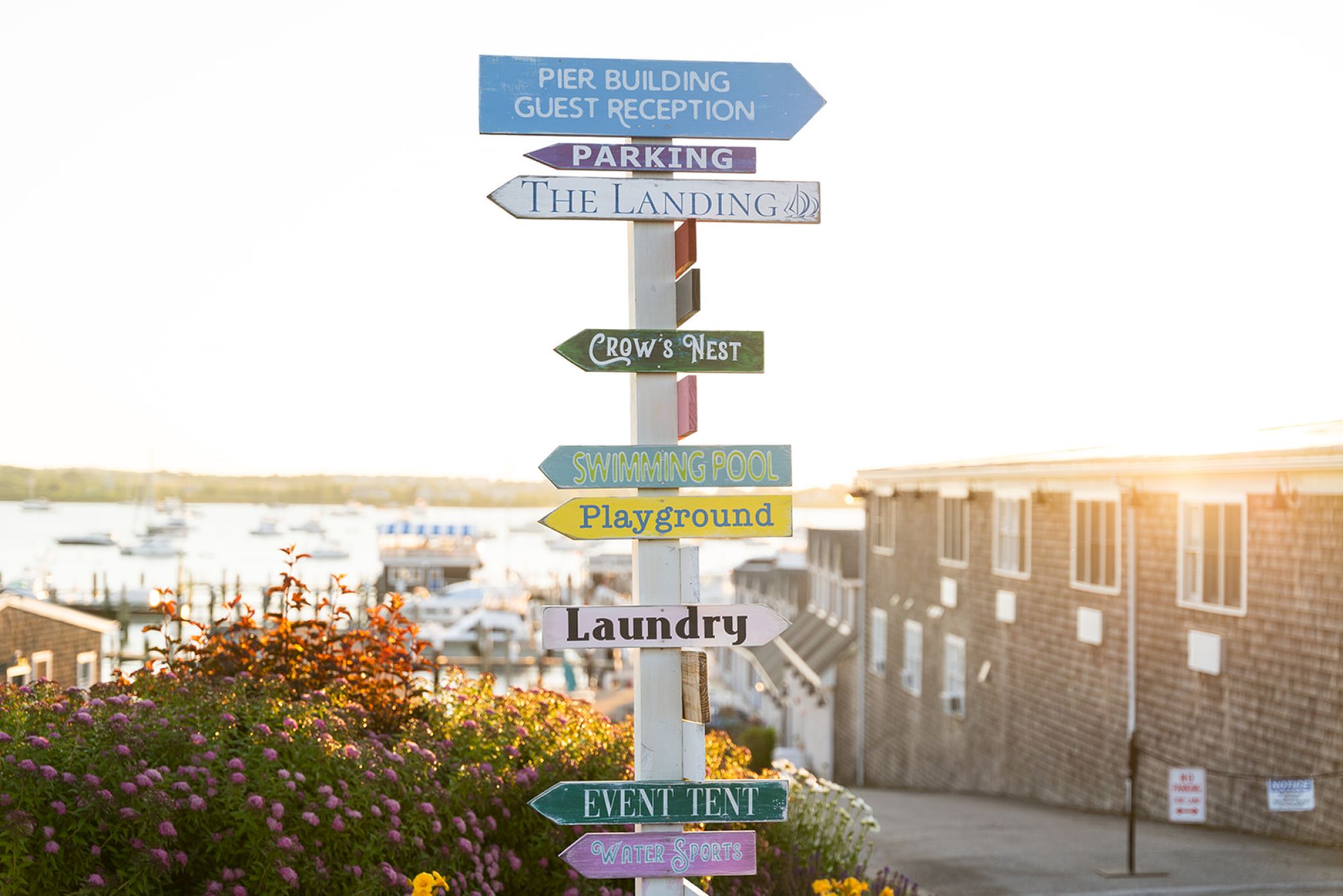 A signpost with multiple colorful directional signs points to various locations like parking, swimming pool, playground, laundry, and event tent.