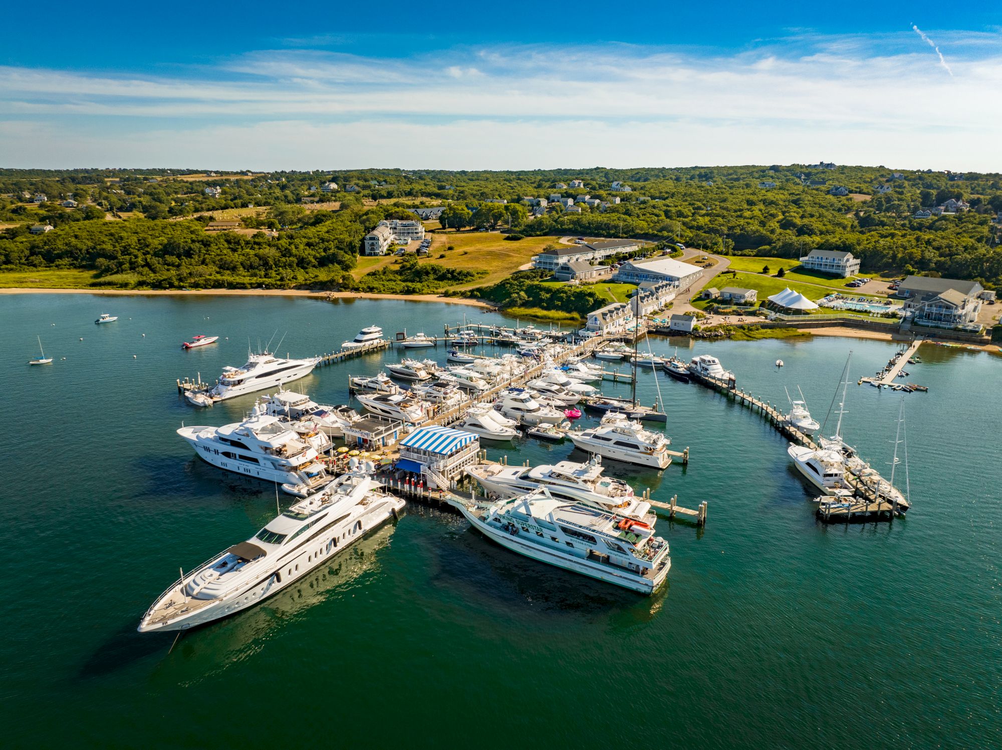 A marina filled with various boats and yachts is visible, with a small town and green landscape in the background under a clear blue sky.