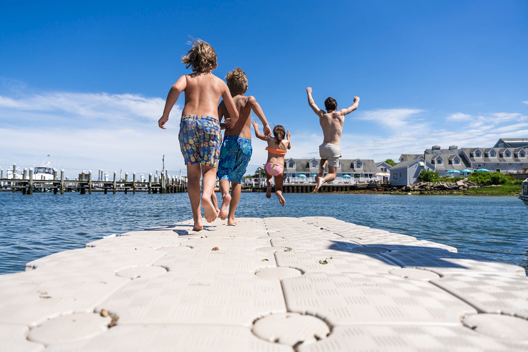 Children running on a dock towards the water, with one jumping in the air, against a backdrop of a marina and buildings under a clear sky.