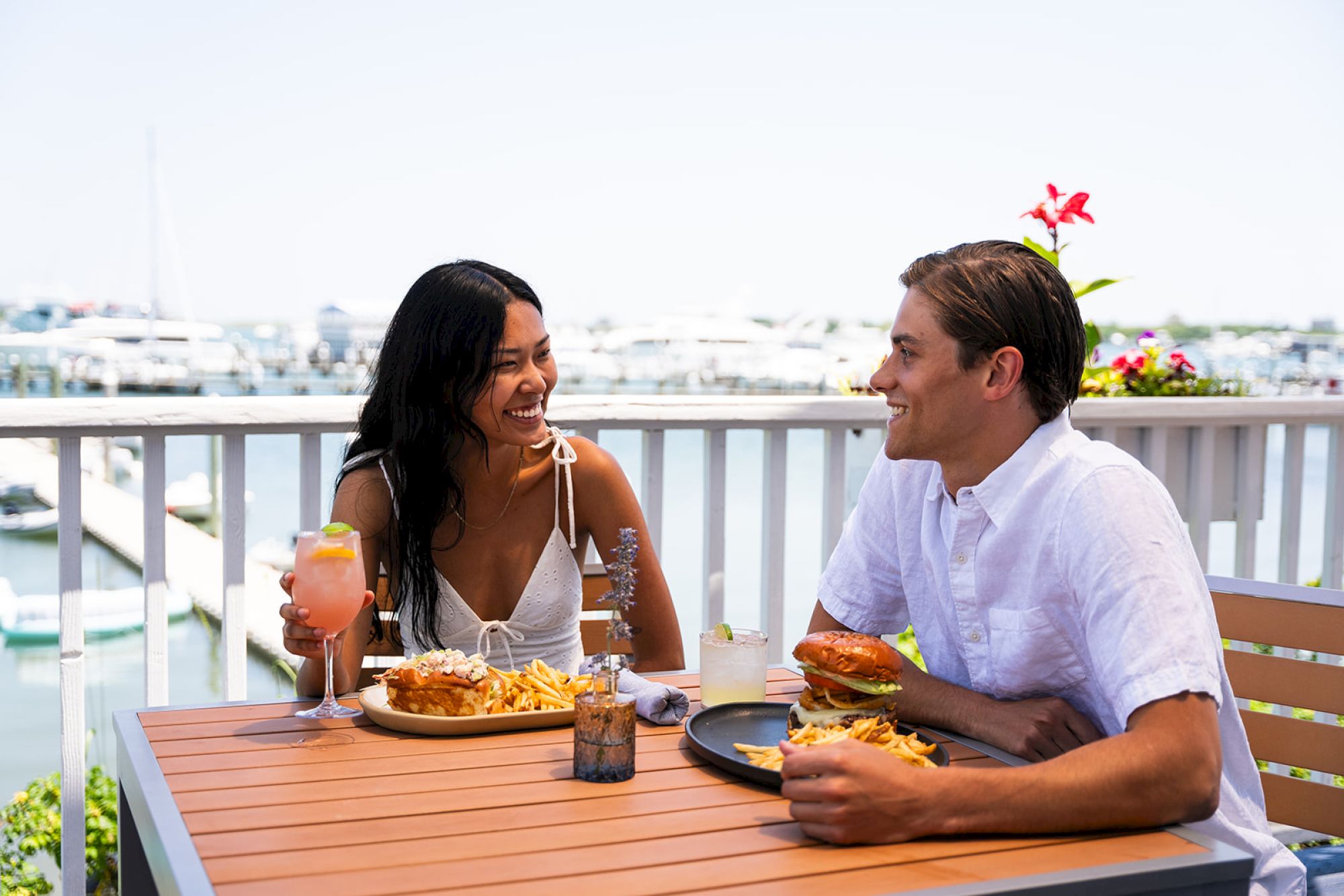 A smiling couple enjoys a meal with fries and drinks at an outdoor seaside restaurant, sitting on a wooden deck with a view of boats in the background.