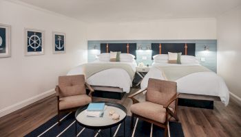 A hotel room with two beds, nautical-themed decor, two chairs, a round coffee table, and wooden flooring. Three framed artworks on the wall.